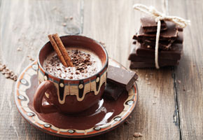 a cup of steaming hot cocoa with a cinnamon stick stirring it with a few pieces of chopped chocolate on a wooden background