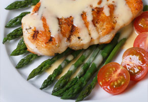 white plate containing grilled chicken, asparagus and sliced tomatoes with avgolemono sauce on a white background