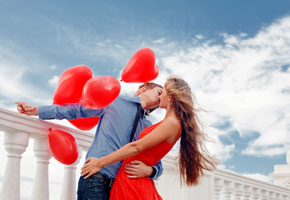 young couple kissing on a pier surrounded by red, heart shaped balloons