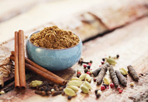 cinnamon, cardamom, nutmeg whole spices and a blue cup containing garam masala on a wooden table
